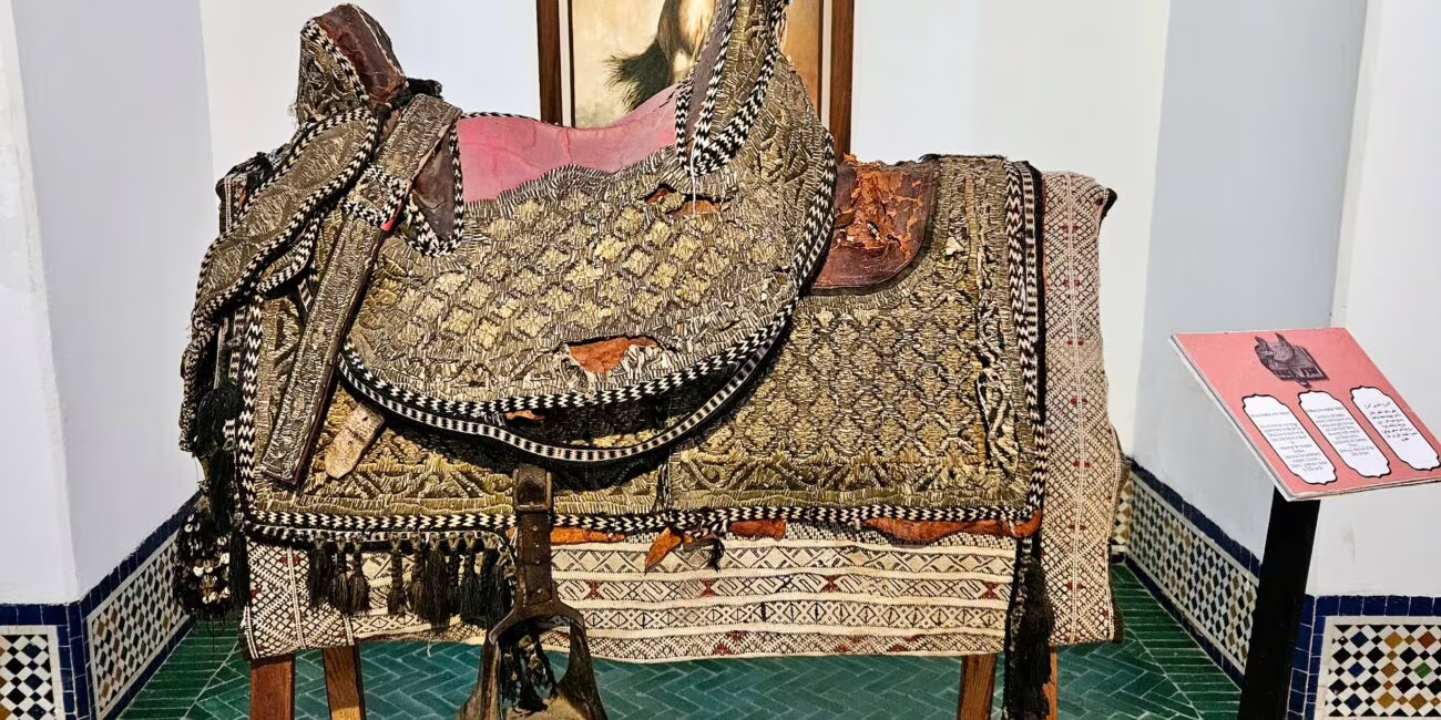 Wystawiona, ozdobna siodło i trek na drewnianym stelażu, pokryte bogatymi tkaninami z fręzlami i ornamentami; za nimi obrazy konia w ramie.