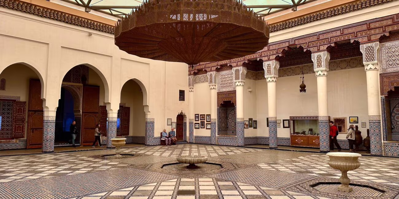 Dworski dziedziniec z mozaikowym podłogowym wzorem, centralnym dużym żelaznym żyrandolem wiszącym nad otwartą przestrzenią, arkadowymi wejściami, kolumnami ozdobionymi kafelkami i ornamentami, oraz grupą zwiedzających przy ścianach.