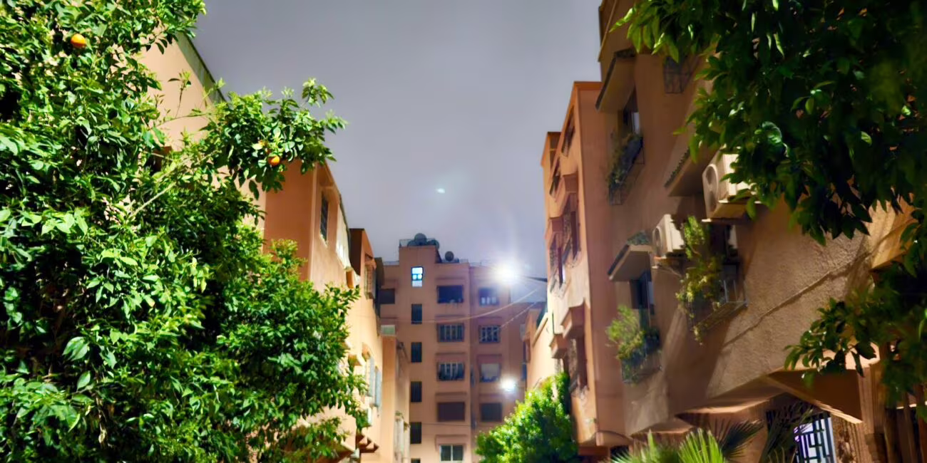 Wąska uliczka wieczorem między wysokimi, pomarańczowymi blokami, otoczona zielenią drzew i palm, z zaparkowanym czarnym samochodem po lewej stronie.