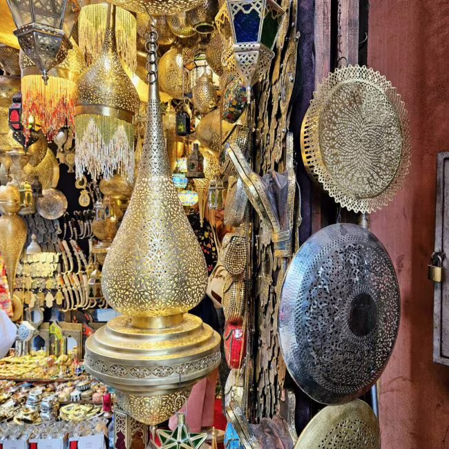 Bogato zdobione metalowe latarnie i misy o skomplikowanych, koronkowych wzorach, wiszące i stojące w tle sklepu z żółtym światłem.