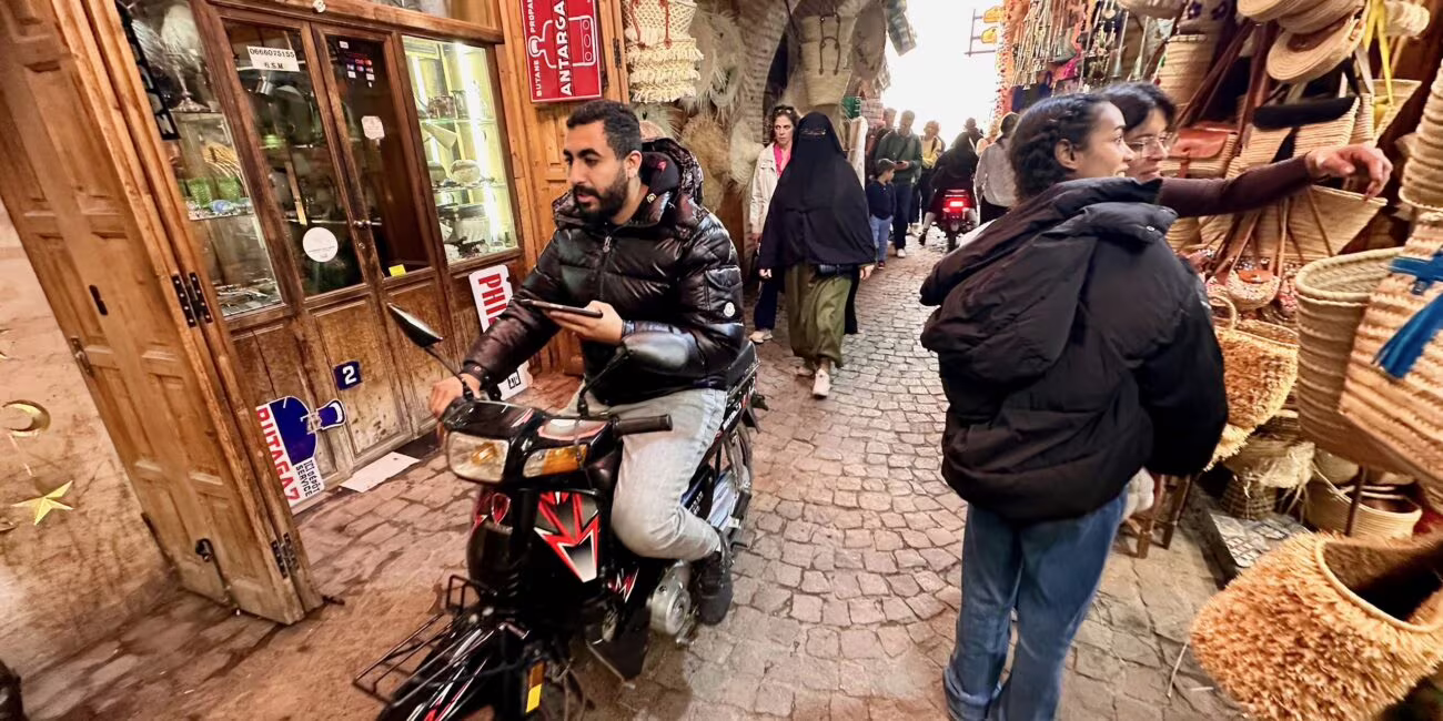 Wąska, kamienna uliczka bazaru z plecionymi koszami i sklepami z drewna; mężczyzna na motorze stoi na ścieżce, kobiety w kurtkach przeglądają towary na wystawach.