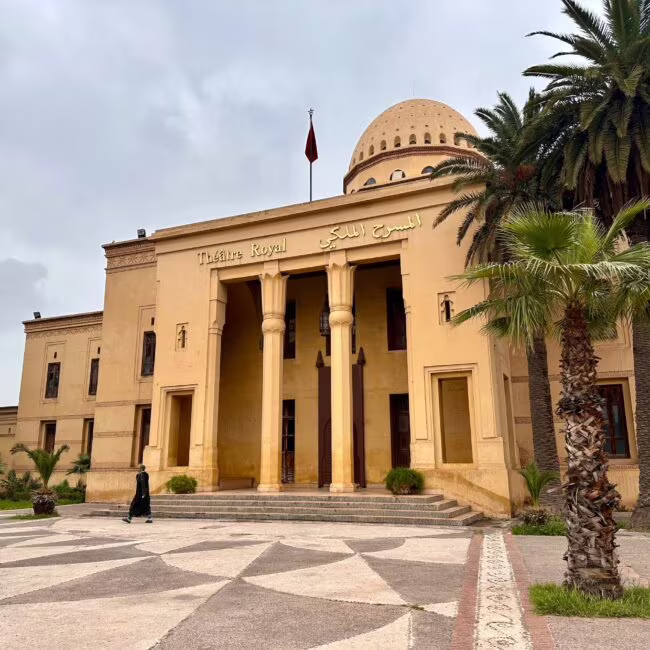 Teatr Royal w budynku w kolorze piaskowym z kolumnami przed wejściem, otoczony palmami i szerokim placem, pomiędzy chmurami.