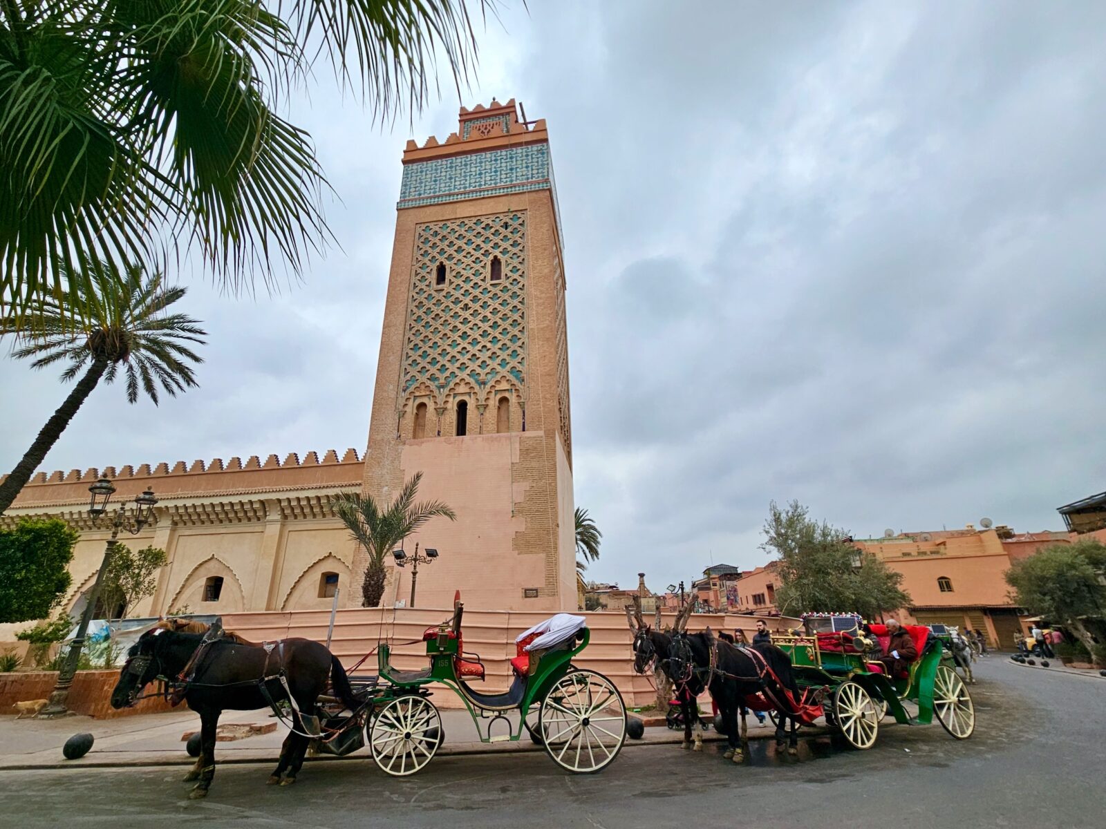 Kawalera konnych karet, zaprzęgane do zielonych piętrowych wozów przed meczetem z wieżą o zdobionej, kolorowej fasadzie i palmami w pobliżu, nadchodzi pochmurny dzień.