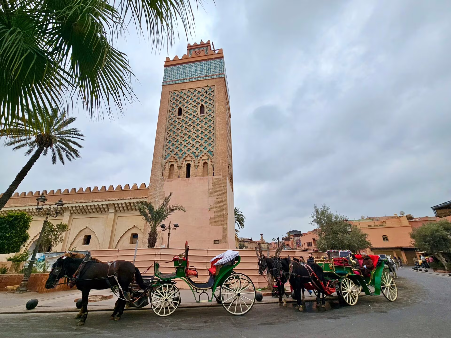 Kawalera konnych karet, zaprzęgane do zielonych piętrowych wozów przed meczetem z wieżą o zdobionej, kolorowej fasadzie i palmami w pobliżu, nadchodzi pochmurny dzień.