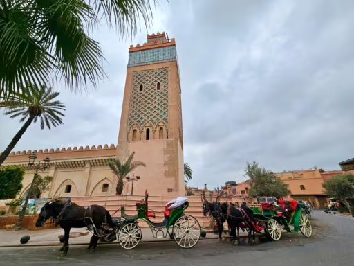 Kawalera konnych karet, zaprzęgane do zielonych piętrowych wozów przed meczetem z wieżą o zdobionej, kolorowej fasadzie i palmami w pobliżu, nadchodzi pochmurny dzień.