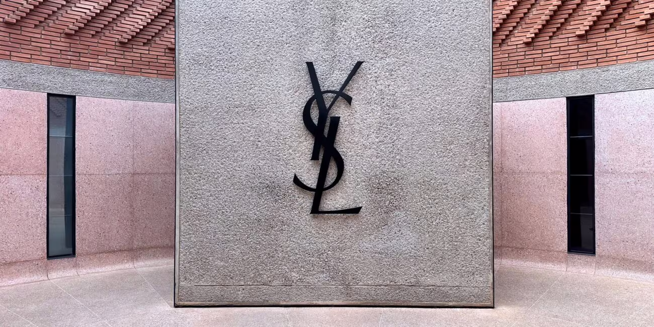 Szara tablica z czarnym monogramem „S”, obok wejścia budynku w ceglanej elewacji.