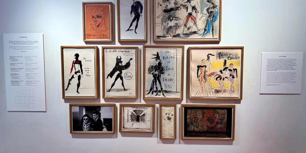 Kolekcja rysunków i plakatów na białej ścianie w galerii, układająca się w prostokątny grid, z różnorodnymi manuskryptami i postaciami w wąskich ramkach.